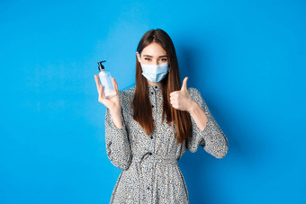 科维德社会距离医疗保健概念美丽的女孩医疗面具手消毒剂持有防腐剂瓶显示拇指站蓝色的背景