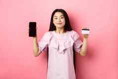 快乐的亚洲女孩显示空智能手机屏幕信贷卡购物在线站粉红色的背景