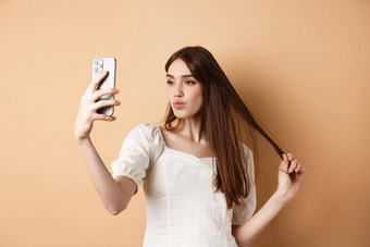 时尚的年轻的女人玩头发采取<strong>自</strong>拍智能手机使照片社会<strong>媒体</strong>站米色背景