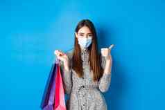 科维德流感大流行生活方式概念有吸引力的女人穿医疗面具购物显示拇指持有袋购买蓝色的背景