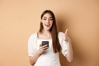 在线购物概念快乐年轻的女人显示拇指手机微笑很高兴站米色背景
