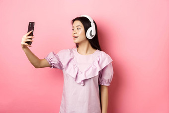 美丽的亚洲女孩博主采取自拍无线耳机使照片社会媒<strong>体智能</strong>手机站衣服粉红色的背景