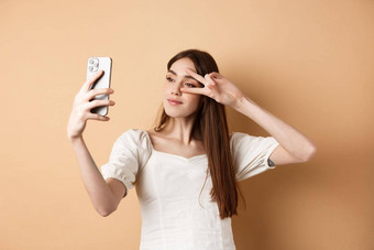 可爱的女人采取自拍智能手机显示地摆眼睛摆姿势社会媒体照片站米色背景