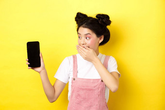 在线购物概念愚蠢的日本女孩美化妆封面口手笑显示空智能手机屏幕显示有趣的事情电话黄色的背景
