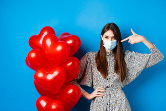 科维德检疫健康问题时髦的年轻的女人医疗面具指出手指红色的心气球皱着眉头失望情人节一天蓝色的背景