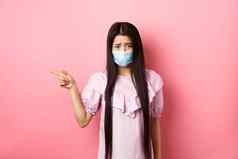 科维德流感大流行生活方式概念伤心悲观的亚洲女孩医疗面具指出左抱怨不公平的情况站失望粉红色的背景