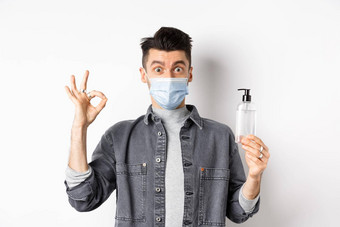 健康的人科维德概念兴奋男人。医疗面具持有瓶好手洗手液显示标志推荐防腐剂站白色背景
