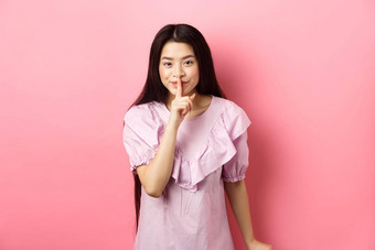 可爱的亚洲女孩隐藏秘密使安静手指按下嘴唇微笑安静的站衣服粉红色的背景