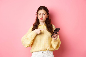持怀疑态度年轻的女人扮鬼脸不满意指出手指智能手机屏幕怀疑在线内容站粉红色的背景