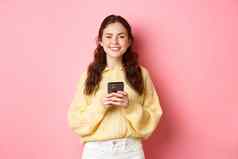 肖像美丽的女孩持有移动电话微笑购物在线社会媒体应用程序站粉红色的背景休闲衣服