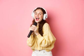 无忧无虑的女孩享受<strong>卡拉ok</strong>晚上唱歌首歌麦克风穿无线耳机站粉红色的背景