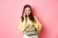 年轻的魅力女孩持有钱包会说话的智能手机微笑休闲谈话电话调用站粉红色的背景