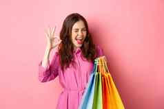 年轻的快乐女人购物者显示标志眨眼很高兴好折扣购买工作人员出售持有购物袋微笑很高兴粉红色的背景