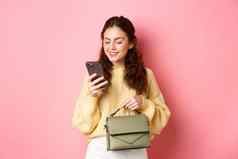魅力女孩持有钱包发短信消息智能手机阅读移动电话屏幕无忧无虑的微笑站粉红色的背景