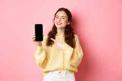 肖像有吸引力的年轻的女人演示了促销电话指出手指智能手机微笑站粉红色的背景