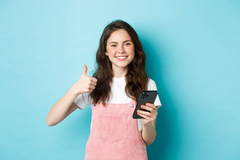 肖像美丽的快乐女孩显示拇指持有智能<strong>手机</strong>在线购物使<strong>订单</strong>互联网满意赞扬好应用程序蓝色的背景