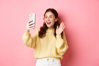 快乐的女孩视频聊天智能手机挥舞着手电话相机微笑快乐站粉红色的背景