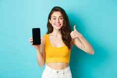 肖像时尚的现代女人微笑很高兴推荐应用程序显示标志横幅空智能手机屏幕使拇指批准赞美应用程序蓝色的背景