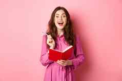 兴奋年轻的女人优秀的的想法写作的想法规划师持有笔记本日记微笑惊讶站粉红色的背景