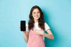 快乐微笑浅黑肤色的女人女孩指出手指空智能手机屏幕推荐在线商店移动应用程序站蓝色的背景