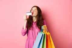 肖像兴奋快乐购物狂女人持有购物袋接吻塑料信贷卡微笑惊讶站粉红色的背景