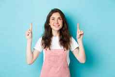 年轻的快乐的女孩魅力使穿粉红色的夏天粗布工作服指出手指微笑促销提供前蓝色的背景