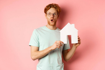 真正的房地产惊讶年轻的男人。红色的头发胡子穿眼镜t恤显示纸房子断路深刻的印象哇站粉红色的背景