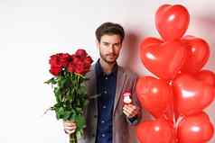 英俊的年轻的男人。准备使建议持有订婚环花束红色的玫瑰使惊喜情人节一天站心气球白色背景