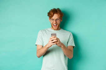 图像年轻的红色头发的人男人。眼镜阅读电话屏幕惊讶脸收到令人惊异的促销提供站绿松石背景