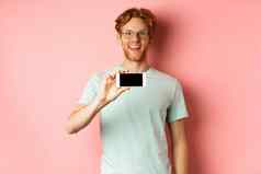 快乐年轻的男人。红色的头发显示智能手机屏幕持有电话水平微笑惊讶站粉红色的背景