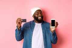 在线购物满意黑色的男人。点头批准微笑电话显示信贷卡智能手机屏幕站粉红色的背景