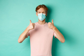 科维德流感大流行生活方式概念快乐的红色头发的人的家伙医疗面具显示拇指批准赞美产品站绿松石背景
