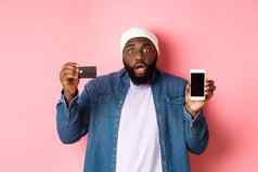 在线购物震惊了有关黑色的男人。盯着相机显示移动电话屏幕信贷卡站粉红色的背景