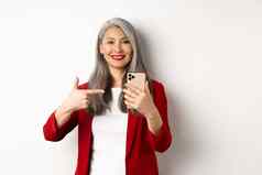 亚洲上了年纪的女人优雅的夹克显示智能手机指出手指移动电话微笑站白色背景