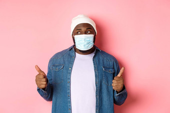 冠状病毒生活方式社会距离概念黑色的赶时髦的人的家伙无檐小便帽医疗面具显示竖起大拇指赞扬产品左微笑粉红色的背景