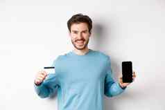 微笑高加索人男人。显示塑料信贷卡移动电话屏幕的家伙推荐在线银行应用程序站白色背景