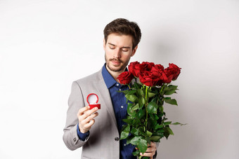 浪漫的男人。西装沉思的订婚环使婚姻建议情人节一天持有花束玫瑰站白色背景
