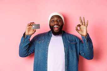 购物概念英俊的非裔美国人男人。推荐银行显示信贷卡标志微笑满意站粉红色的背景