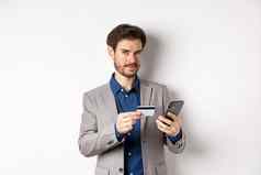 在线购物英俊的商人西装支付信贷卡智能手机微笑满意相机站白色背景