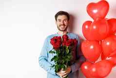 情人节一天浪漫兴奋年轻的男人。花束红色的玫瑰心气球微笑相机带礼物情人情人节日期站白色背景