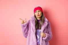 时尚购物概念持怀疑态度中年亚洲女人紫色的皮毛外套指出手指左不高兴伤心表达式站粉红色的背景
