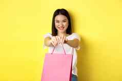 可爱的亚洲女孩给礼物拉伸手粉红色的购物袋微笑祝贺假期站黄色的背景