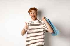 快乐年轻的男人。红色的头发购物商店显示竖起大拇指持有纸袋肩膀推荐商店白色背景
