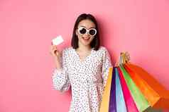 美丽的亚洲女人太阳镜购物持有袋显示信贷卡站粉红色的背景