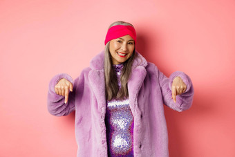 时尚购物概念微笑高级亚洲女人指出手指显示商店广告底穿时尚的紫色的冬天外套粉红色的背景