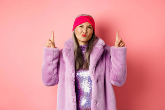 时尚的中间岁的亚洲女人紫色的假的皮毛外套持怀疑态度扮鬼脸不高兴脸指出手指坏不惹人注意的粉红色的背景