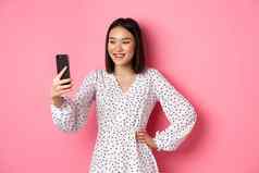 美丽的亚洲女孩照片过滤器应用程序采取自拍智能手机摆姿势可爱的衣服粉红色的背景