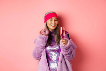 优雅的时尚的亚洲女人紫色的人造皮毛外套吃甜甜圈喝香槟有趣的聚会，派对站粉红色的背景