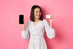 快乐年轻的亚洲女人互联网购物显示智能手机屏幕满意信贷卡站粉红色的背景