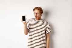 技术电子商务概念惊讶震惊了红色头发的人的家伙检查在线促销活动显示空白智能手机屏幕下降下巴白色背景